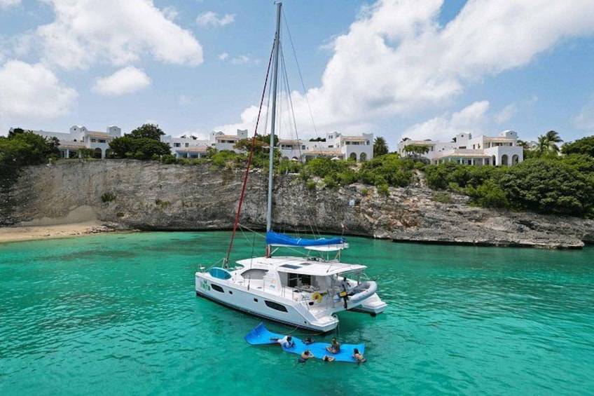 A white sailboat with a blue canopy anchored near a rocky shoreline with white buildings in the background. 2