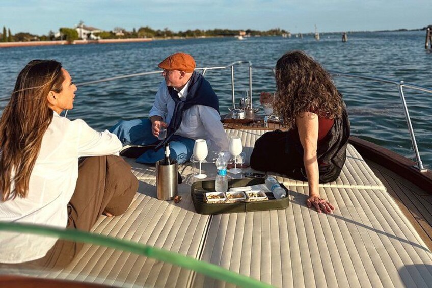 Un bote con tres personas sentadas en la cubierta, una mesa con bebidas y aperitivos, y un cuerpo de agua tranquilo al fondo. 5