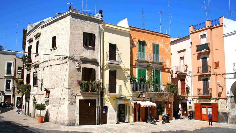 A street with colorful buildings, a shop named 'MACELLERIA,' and a person standing near the entrance. 3