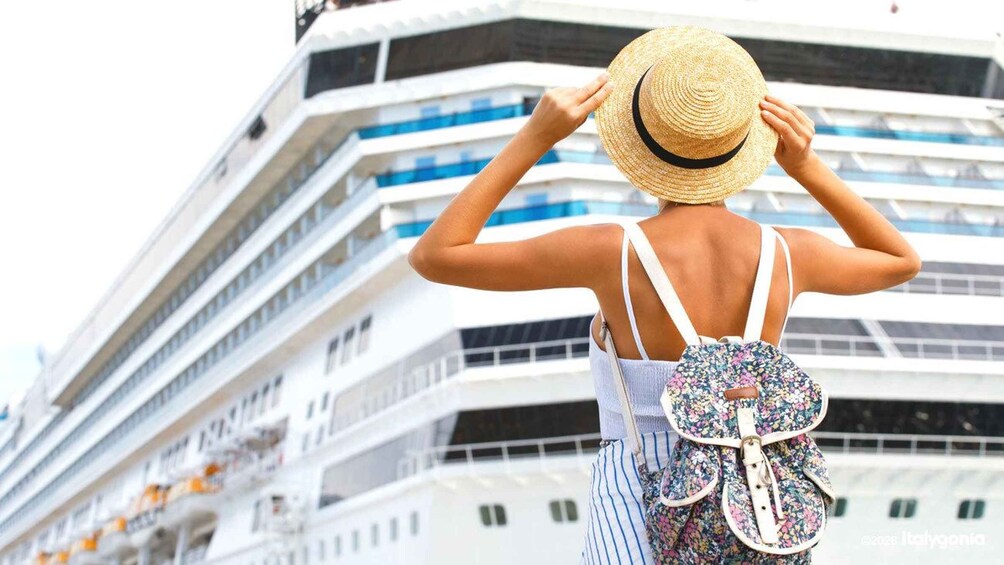 A person wearing a straw hat and a floral backpack, standing in front of a cruise ship. 1