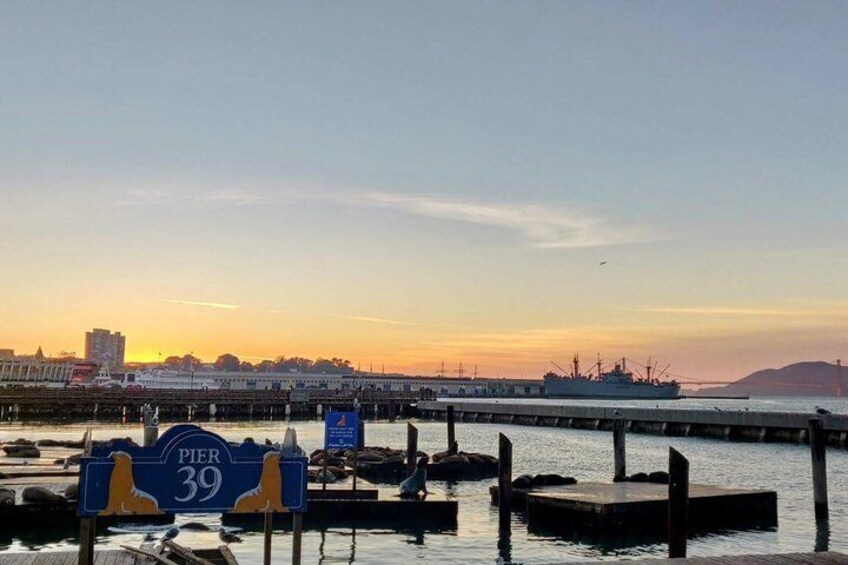 En solnedgång vid Pier 39, med ett stort fartyg förtöjt, en brygga som sträcker sig ut i vattnet och en stadsbild i bakgrunden. 4