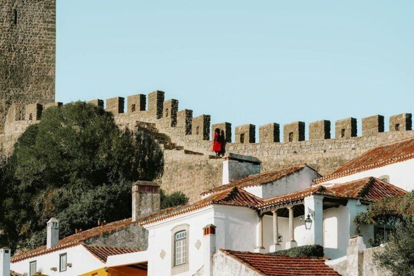 A historic stone wall with a tower, white buildings with red-tiled roofs, and a person in red clothing. 2