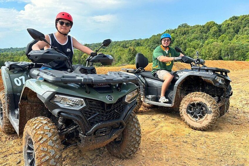 Two people on all-terrain vehicles in a dirt field with trees in the background. 2
