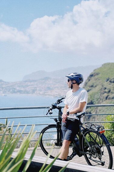 A person standing next to a bicycle on a coastal path with a view of the sea and hills in the background. 5