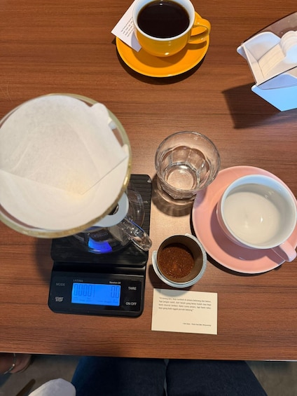 A coffee brewing setup with a scale, a cup of coffee, a glass of water, a bowl of coffee grounds, and a cup of milk on a wooden table. 4