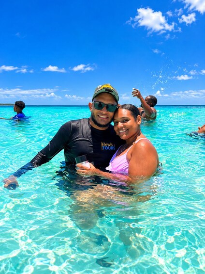Two people in clear turquoise water, a man in a black wetsuit and a woman in a pink swimsuit, with a clear blue sky and distant horizon. 2
