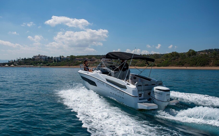 A white motorboat with a black canopy, labeled "LATCHI WATERSPORTS," moving on a body of water with a clear sky and a distant shoreline. 1