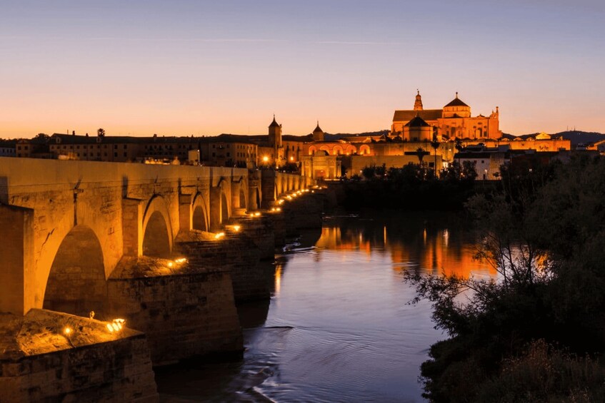 A bridge with arches at dusk, a river, and a large building with domes in the background. 1