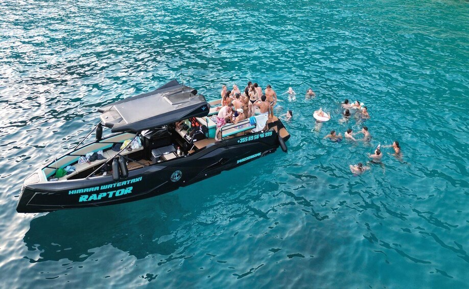 A black boat named 'Himara Water Taxi Raptor' with people swimming and floating around it on clear blue water. 1