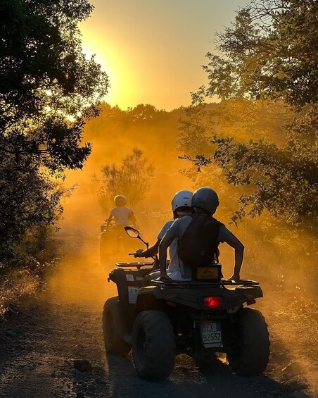 Two people on an ATV on a dusty road with trees on either side, during sunset. 1