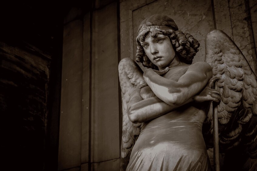 A marble statue of an angel with wings, holding a staff, and standing against a wall. 1