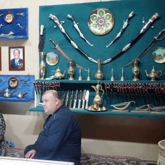 A man sitting in front of a display of antique knives, swords, and decorative items on a teal wall. 4