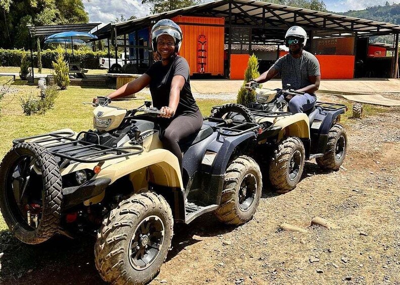 Two people on all-terrain vehicles (ATVs) parked on a dirt path with a building and trees in the background. 1