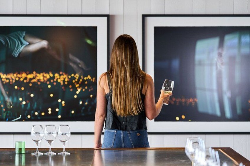 Eine Person hält ein Weinglas und steht vor einer Bar mit Gläsern auf der Theke, während sich zwei große gerahmte Bilder an der Wand befinden. 1