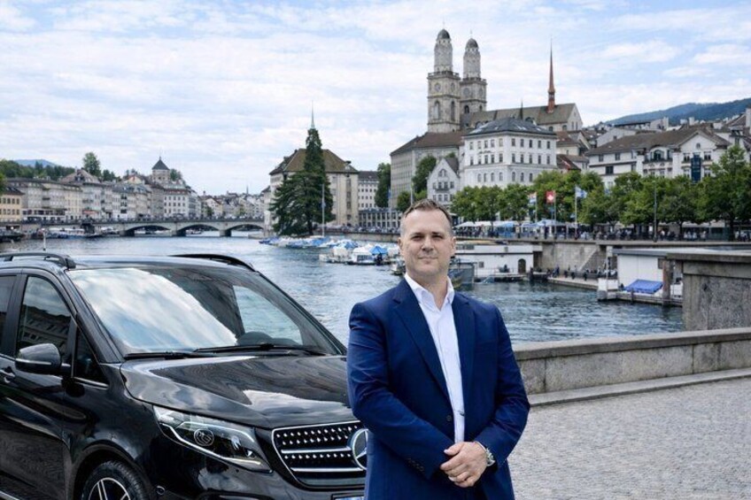 Un hombre con traje azul de pie junto a un coche negro, con un río y edificios históricos al fondo. 1
