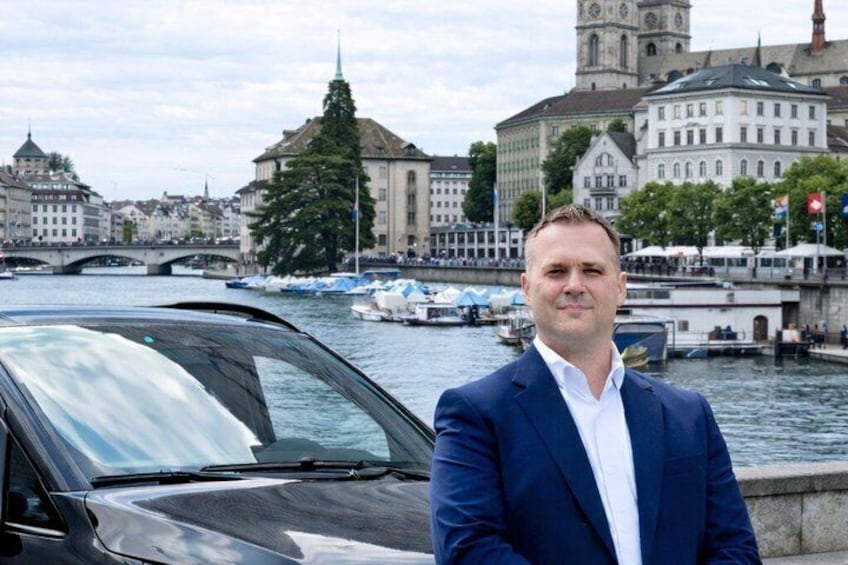 Un hombre con traje azul de pie junto a un coche, con un río y edificios históricos al fondo. 2