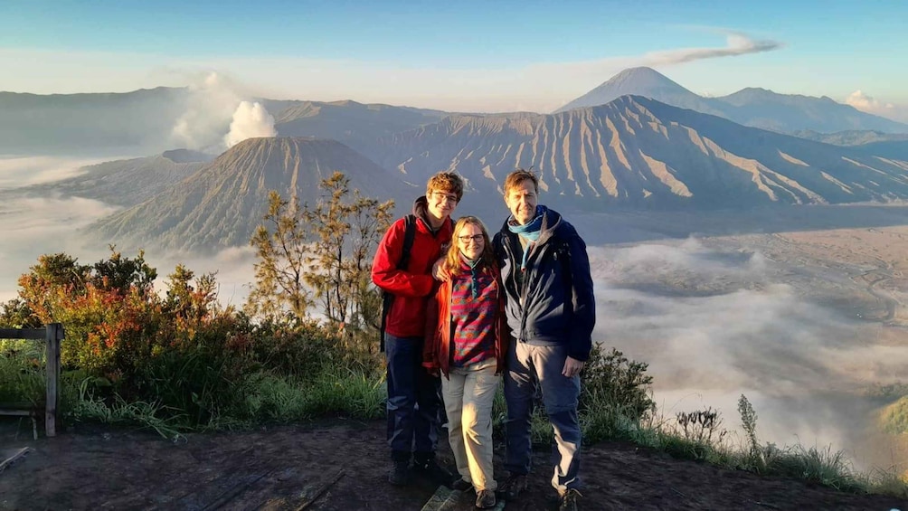 Three people standing on a wooden platform with a mountain and a volcano in the background. 1