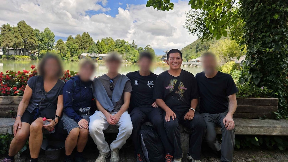 A group of people sitting on a bench in a park with a lake and trees in the background. 2