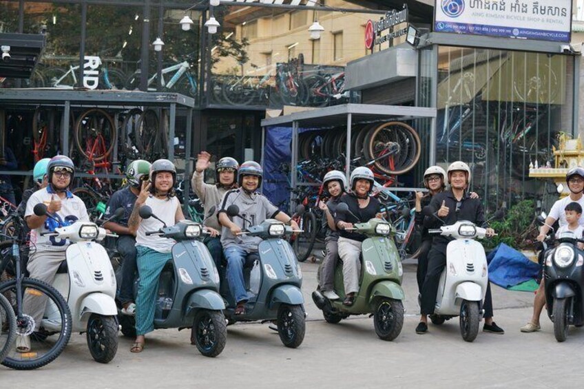 A group of people on scooters in front of a building with bicycles. 2