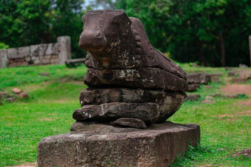 A stone sculpture of a lion's head on a pedestal in a grassy area with trees in the background. 3