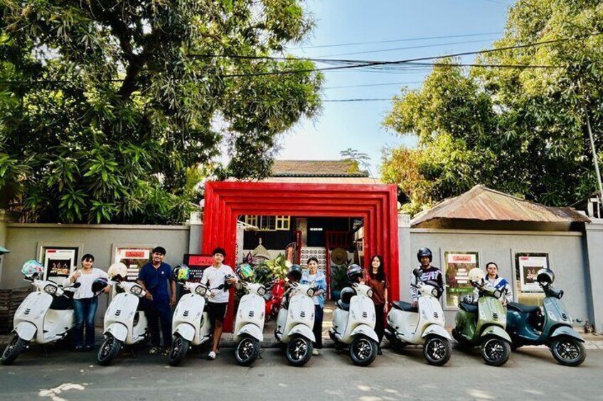 A group of people standing in front of a red building with scooters lined up in front. 1