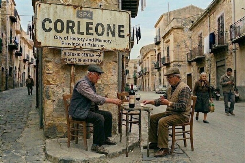 Deux hommes sont assis à une table dans une rue, devant une enseigne indiquant : « Corleone, ville d’histoire et de mémoire, province de Palerme ». 1