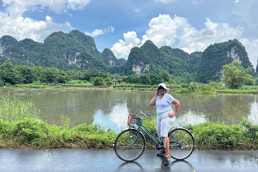 Una persona de pie junto a una bicicleta sobre un camino pavimentado, cerca de una masa de agua tranquila, con vegetación verde y montañas al fondo. 1