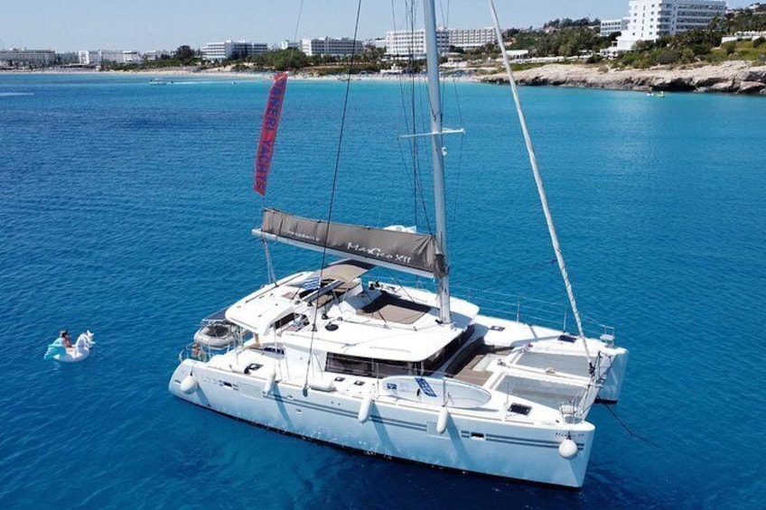A white sailboat with a cabin and a red flag on the mast, anchored in clear blue water with a beach and buildings in the background. 1