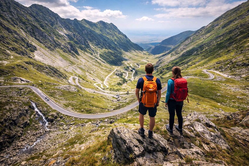 Two people standing on a rocky outcrop overlooking a winding mountain road. 1