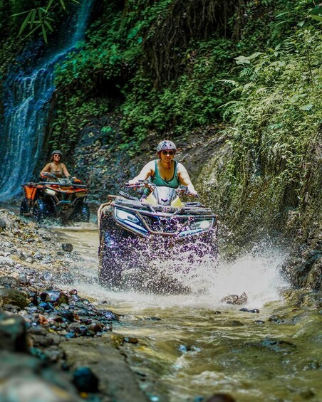 Two people riding ATVs through a shallow river surrounded by dense forest. 1