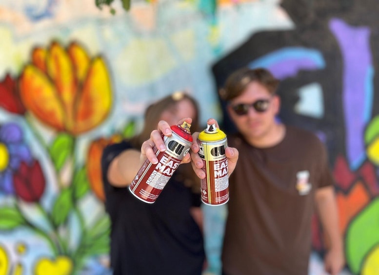 Two people holding spray paint cans in front of a colorful mural. 1