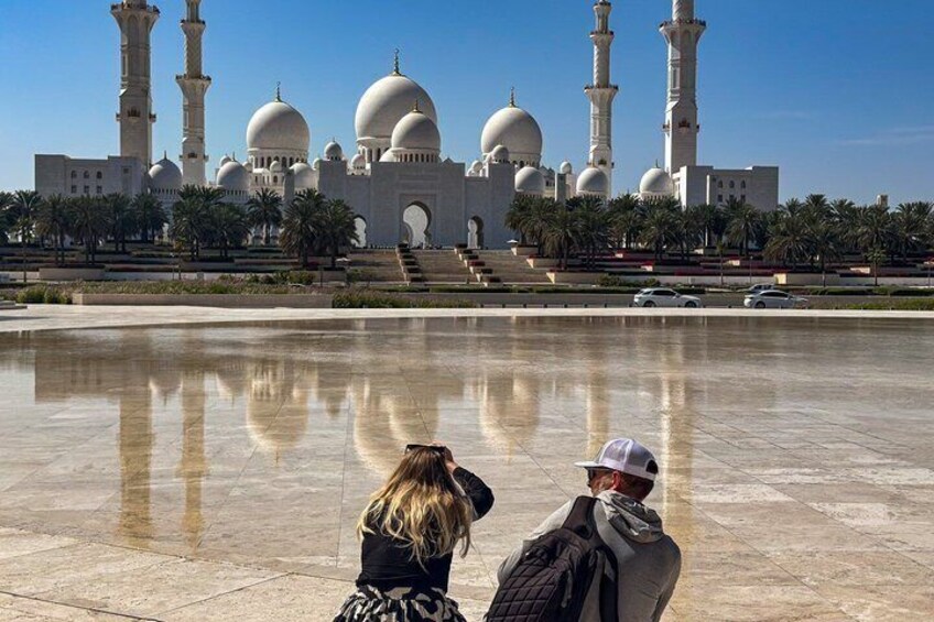 A large mosque with multiple domes and minarets, palm trees, and a reflective water surface. 1