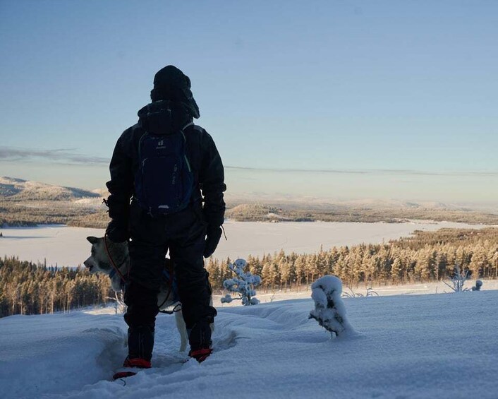 A person with a dog on a snowy mountain overlooking a forest and a lake. 1