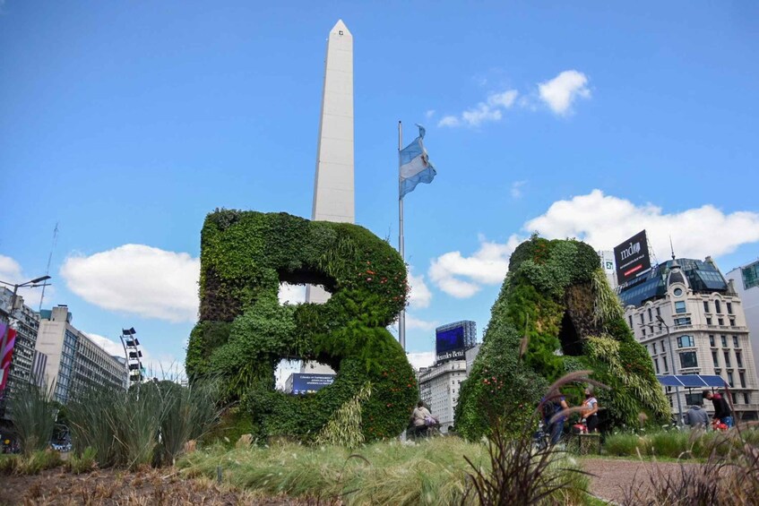 A large obelisk with a flag, surrounded by greenery and urban buildings. 3