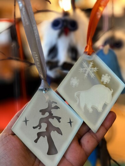 Two square ornaments with tree and snowflake designs, held by a hand. 2