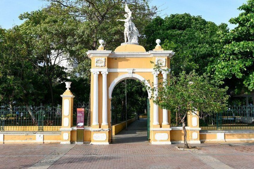 A yellow archway with a statue on top, surrounded by trees and a fence. 1