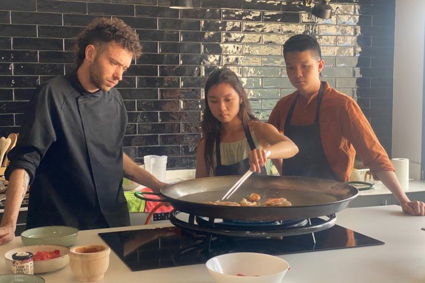 Drei Personen kochen in einer modernen Küche mit schwarzer Ziegelwand und einem großen Wok auf einem Induktionskochfeld. 4
