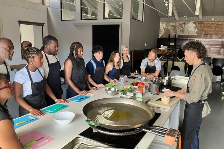 En gruppe mennesker i et moderne køkken, nogle bærer forklæder, og der er køkkenredskaber og ingredienser på bordet. 5
