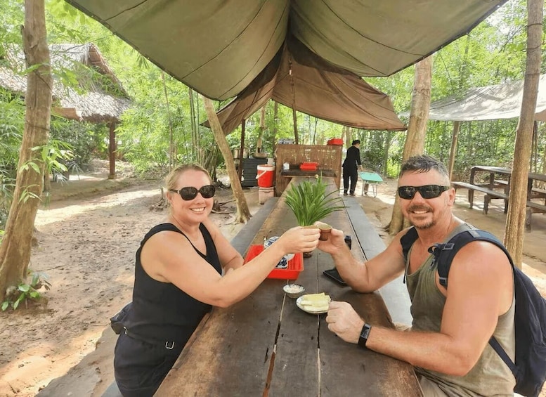 Two people toasting at a wooden picnic table under a canopy in a forested area. 1