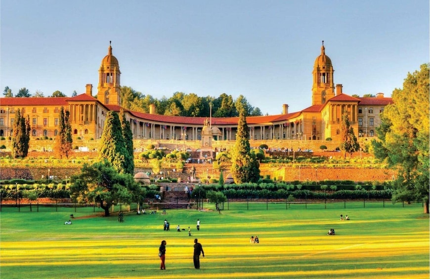 A large, historic building with two towers, surrounded by a well-maintained park with people walking and sitting on the grass. 2