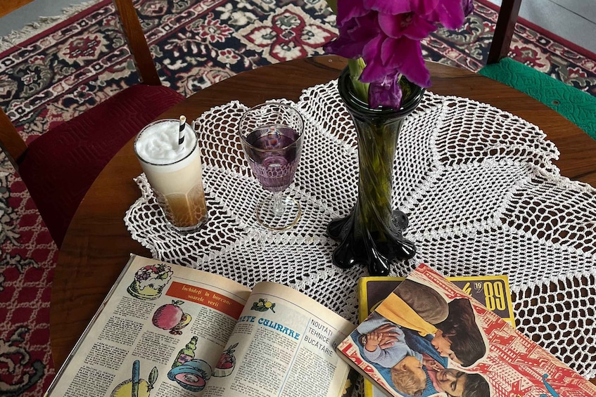 A table with a lace tablecloth, a glass of milk, a vase with purple flowers, and a magazine. 3