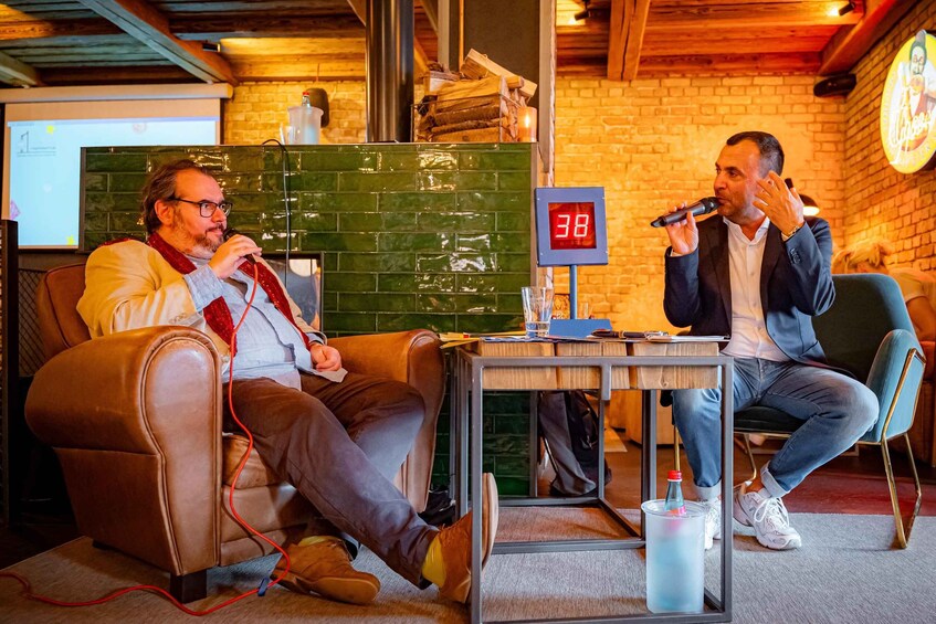 Two men in a room with a brick wall, a green tiled partition, and a digital clock displaying '38'. 3