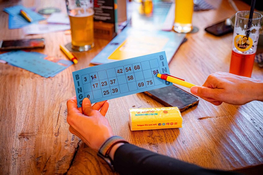A person holding a bingo card with numbers, a smartphone, and a yellow game piece on a wooden table. 1