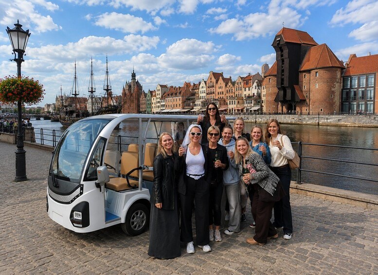 A group of people posing in front of a modern vehicle with a river and historic buildings in the background. 5