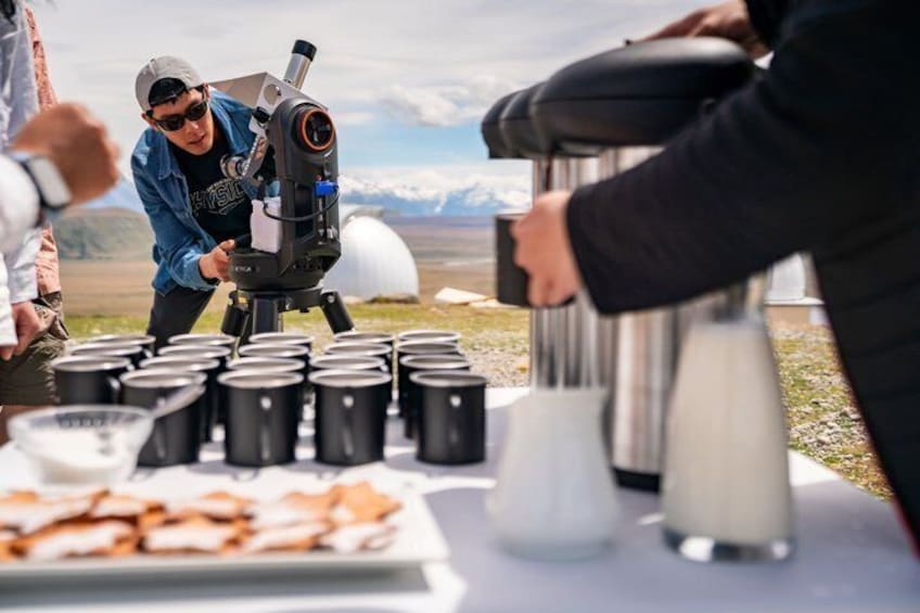 Un télescope posé sur un trépied, des personnes assises autour d’une table avec de la nourriture et des boissons, et un paysage montagneux en arrière-plan. 3