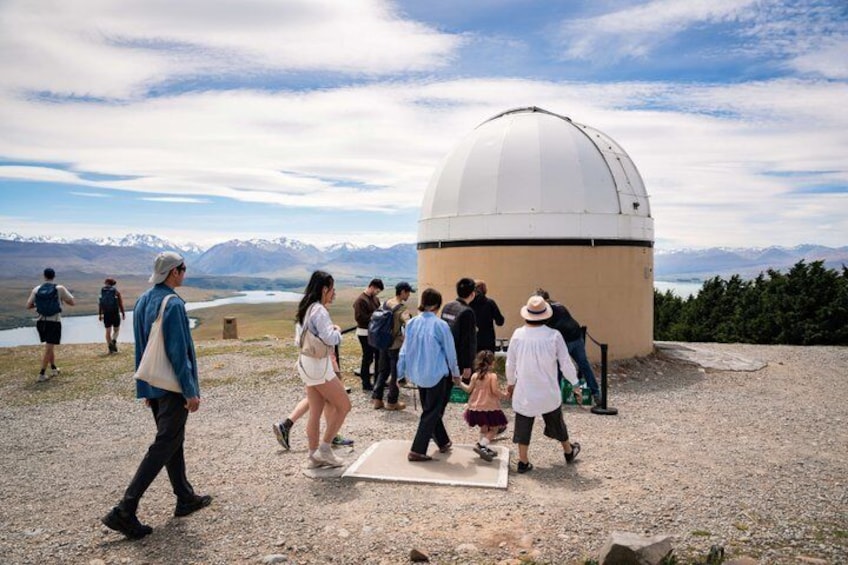 Un groupe de personnes marchant en direction d’un grand dôme d’observatoire situé dans une région montagneuse, avec un lac en arrière-plan. 2