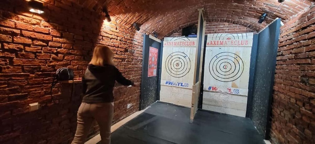 A person aiming at a target in a brick-walled room with two targets labeled "AXEMATECLUB". 2