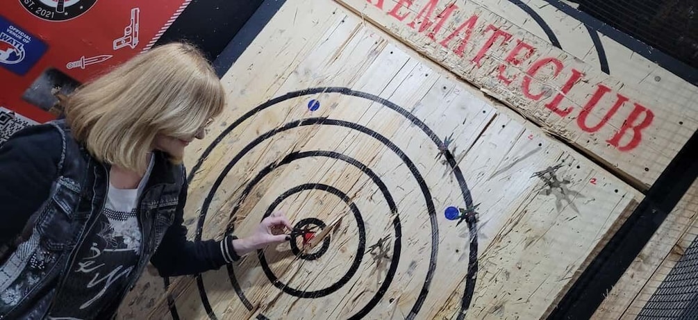 A person aiming at a target with concentric circles on a wooden board with the text "REMAKECLUB" visible. 5