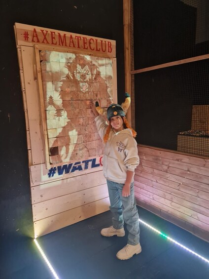 A person in a #AXEMATECLUB hoodie and a hat with horns, standing in front of a wooden structure with a #WATL logo. 4