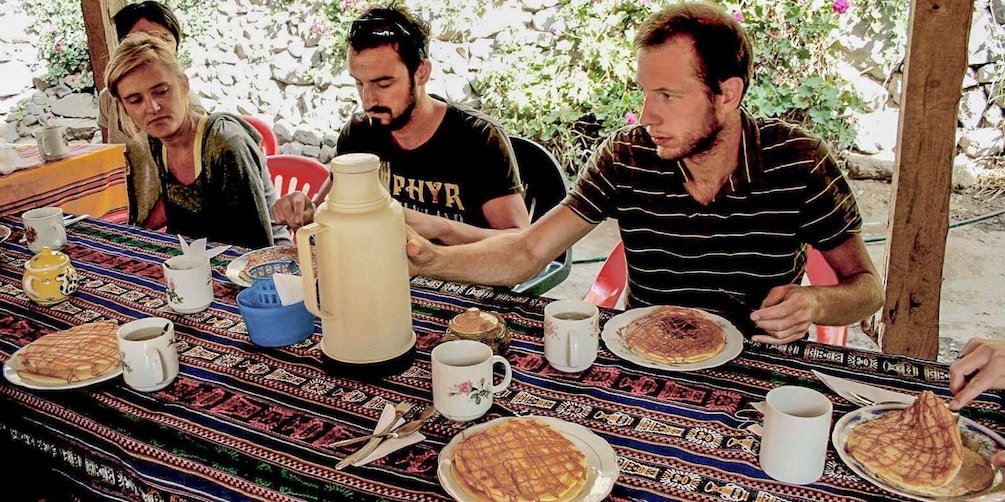 People sitting at a table with food and drinks, outdoors with greenery in the background. 1